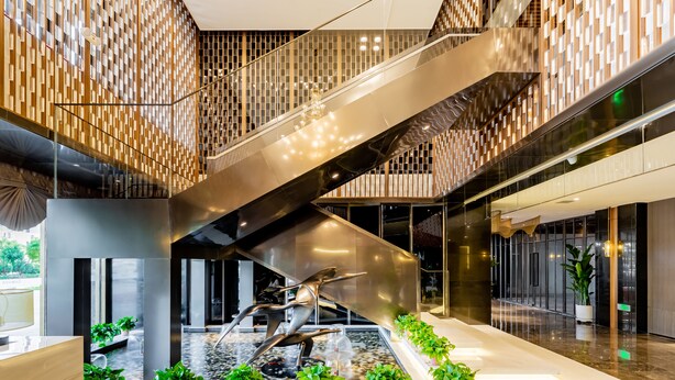 Un interior moderno con una escalera central, barandillas de vidrio y una fuente con plantas.