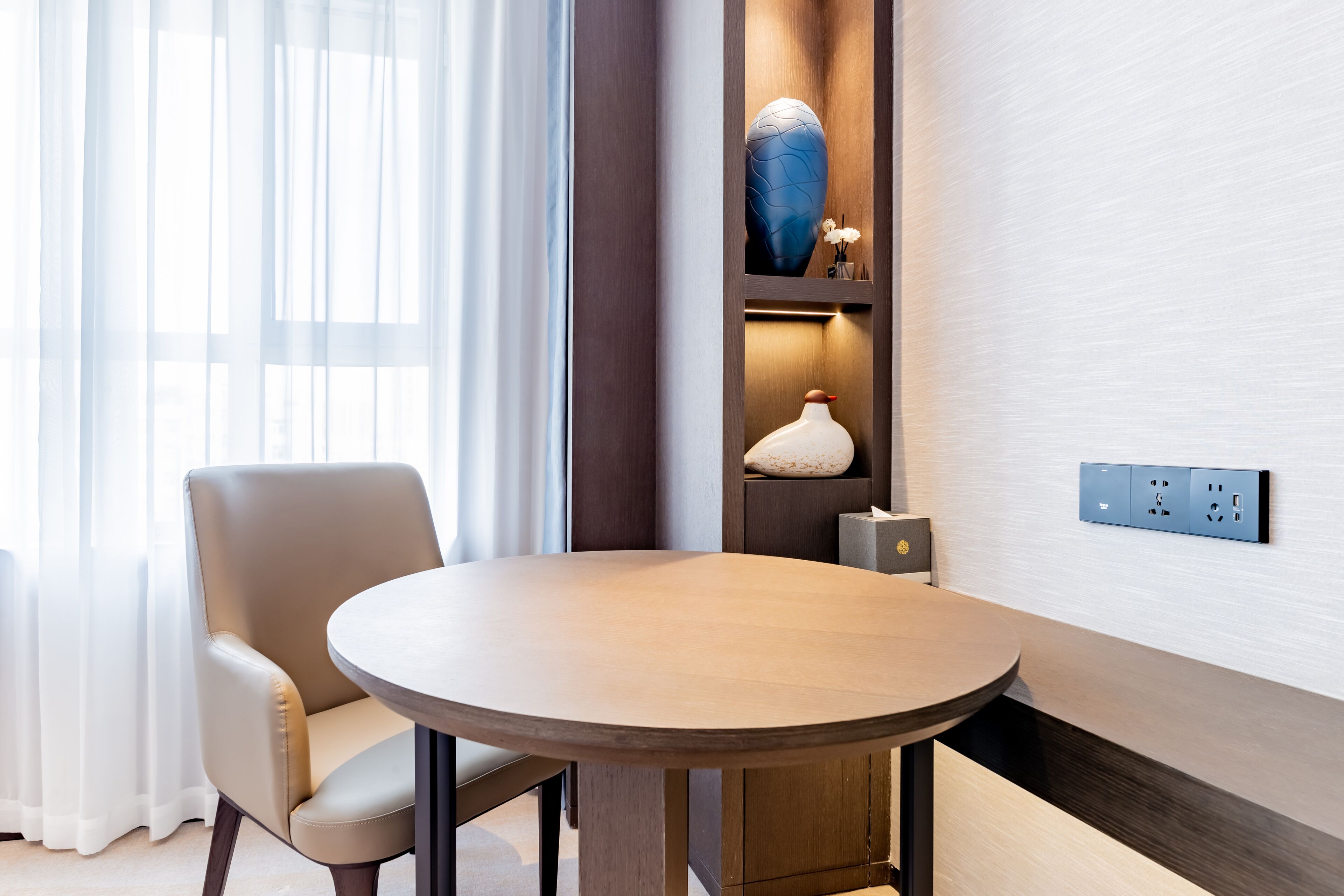 A modern hotel room with a round wooden table, a comfortable chair, a decorative blue vase, and a wall-mounted electrical outlet.