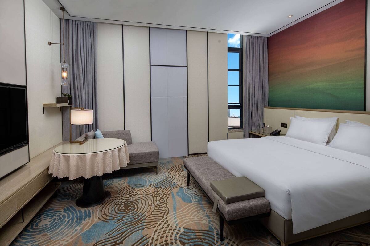 A hotel room with a large bed, a small round table, a sofa, a TV, and a view of the cityscape.