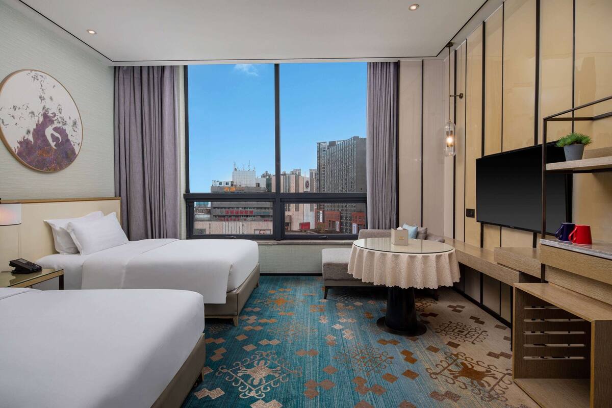 A hotel room with two beds, a round table, a TV, and a city view.
