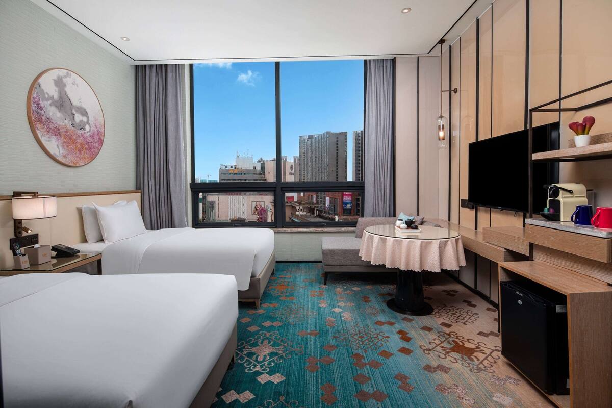 A hotel room with two beds, a TV, a small table with a tablecloth, a desk, and a view of the city through large windows.