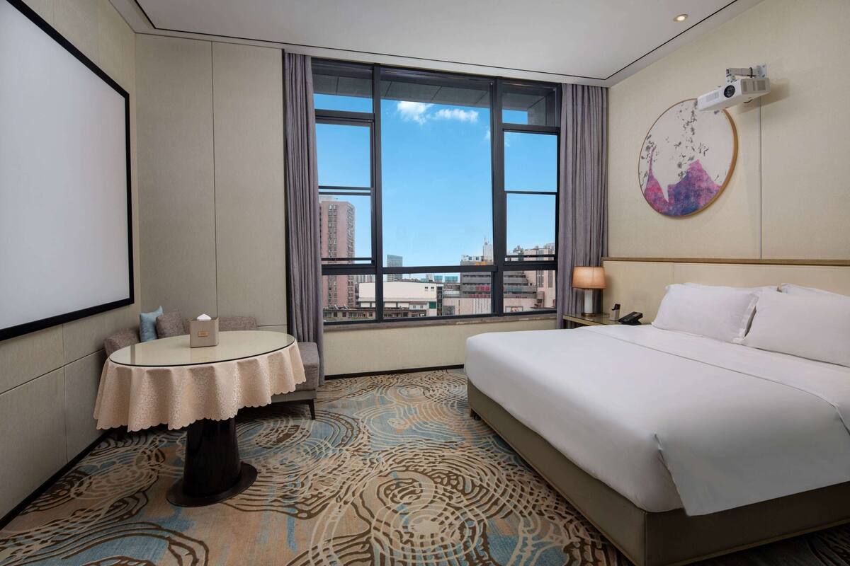 A hotel room with a large bed, a round table with a lamp, a projector on the wall, and a view of the city through large windows.