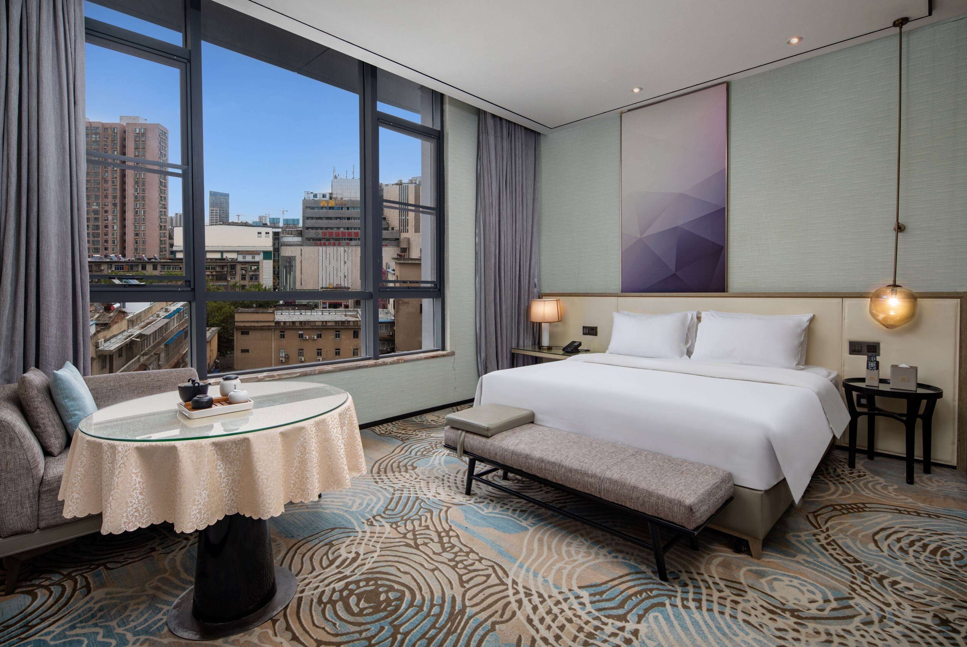 A hotel room with a large window, a bed, a sofa, a small table, and a bench.