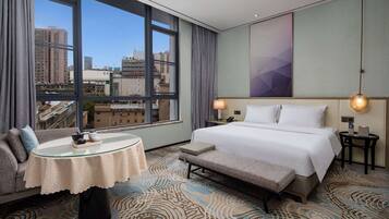 A hotel room with a large window, a bed, a sofa, a small table, and a bench.