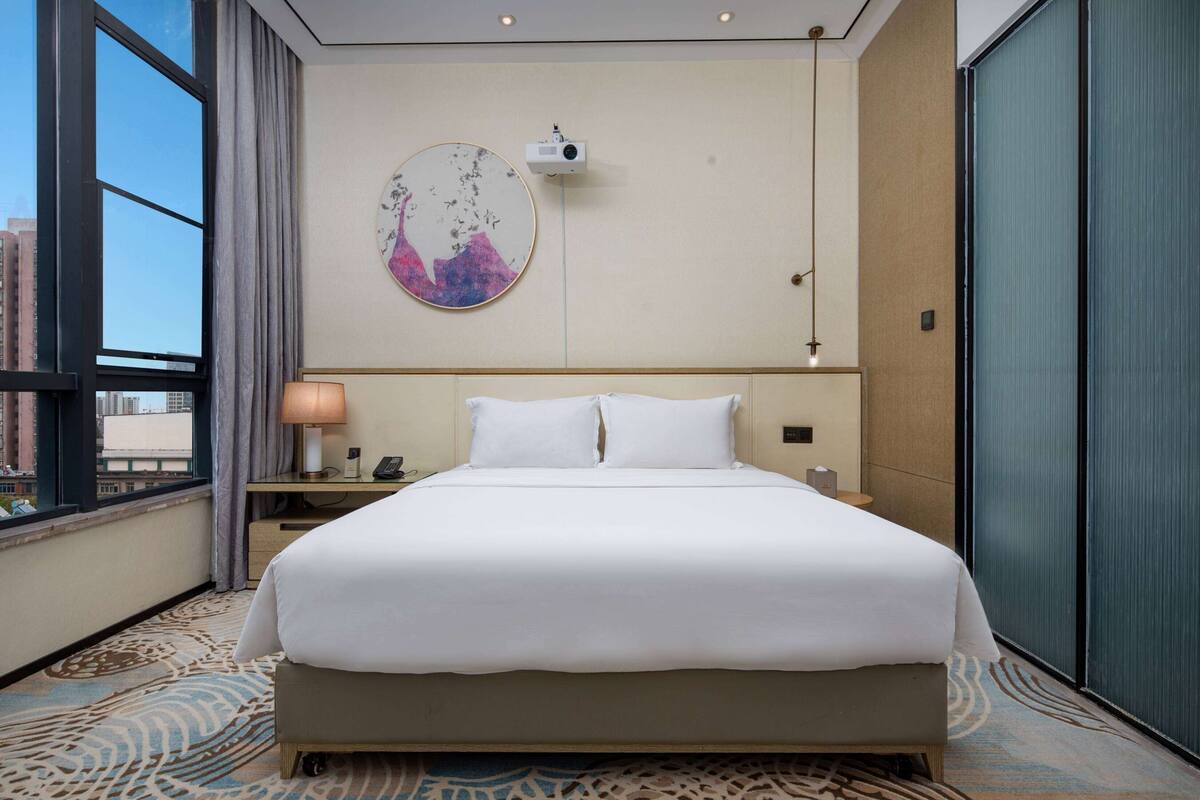 A hotel room with a large bed, a desk with a lamp, a phone, a wall-mounted projector, and a large window with a view of buildings.
