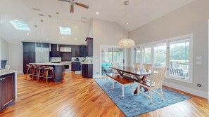 A spacious kitchen with dark wood cabinets, a central island with stools, a dining area with a large table and chairs, and large windows offering views of the outdoors.