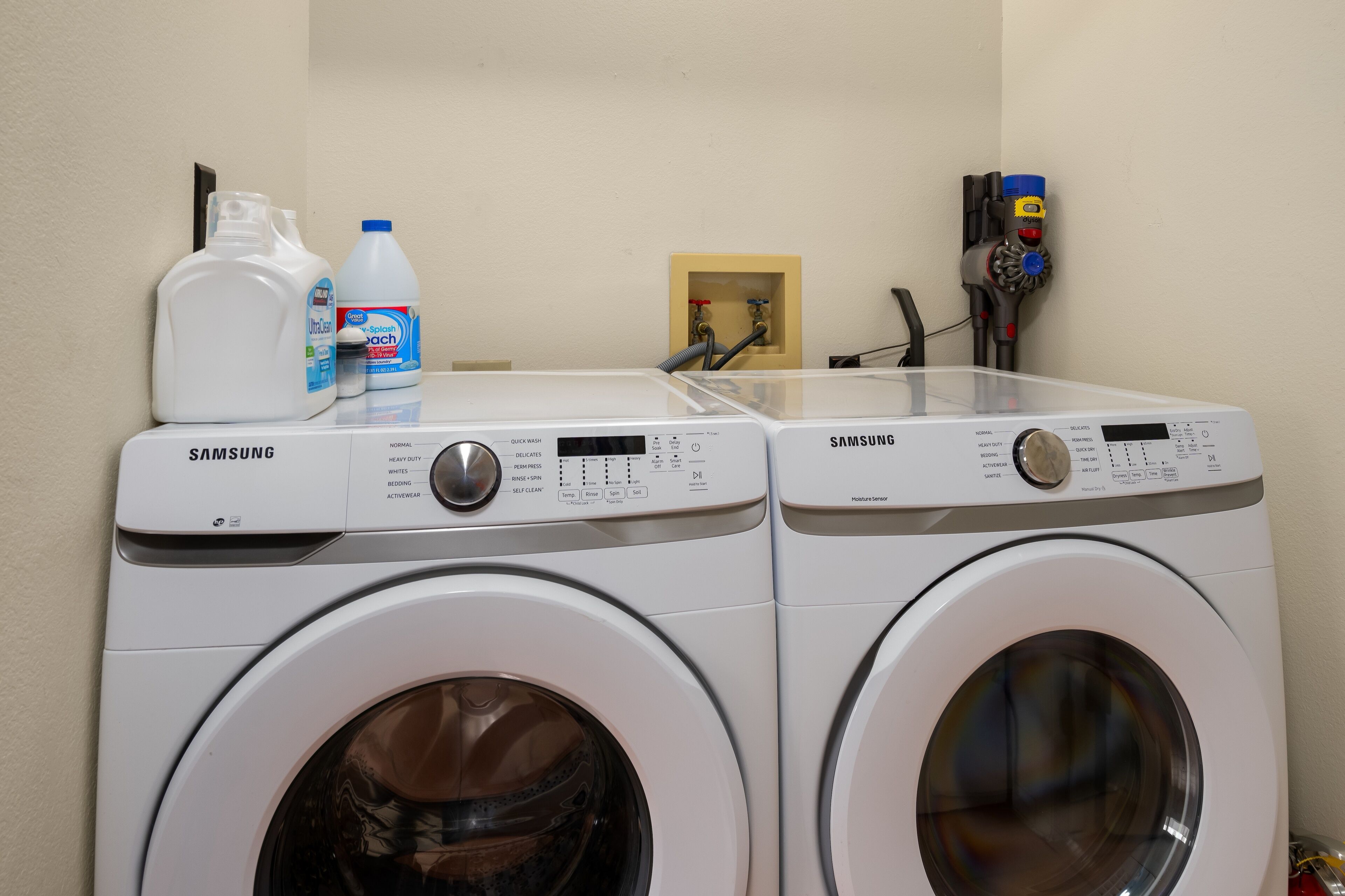 Two Samsung front-loading washing machines in a laundry area with a water heater and detergent bottles on the counter.