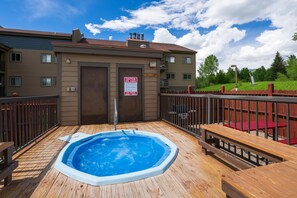 A wooden deck with a hot tub, a sign indicating spa hours, and a building with a brown facade and multiple windows.