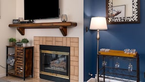 A living room with a flat-screen TV, a fireplace, a wooden shelf, a side table with a lamp, and a wall-mounted mirror.