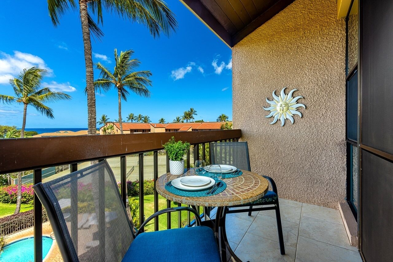 Un balcón con una mesa dispuesta para dos personas, con vistas a una piscina y palmeras.