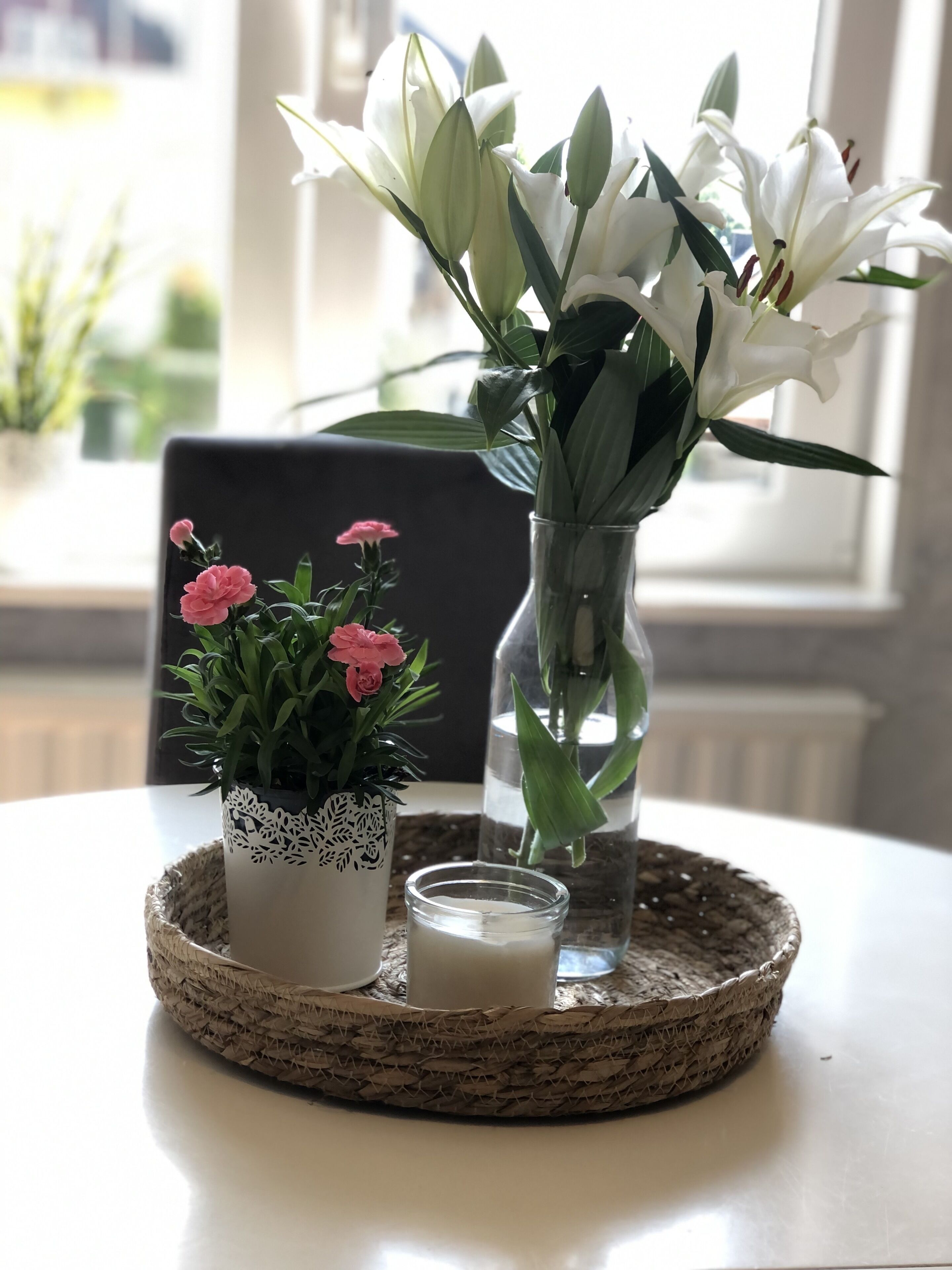 A table with a woven tray holding a potted plant with pink flowers, a clear glass vase with white flowers, and a candle.