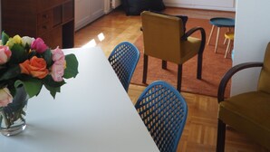Una sala de estar con una mesa blanca, sillas azules, un sofá, un televisor en la pared y una ventana con persianas.