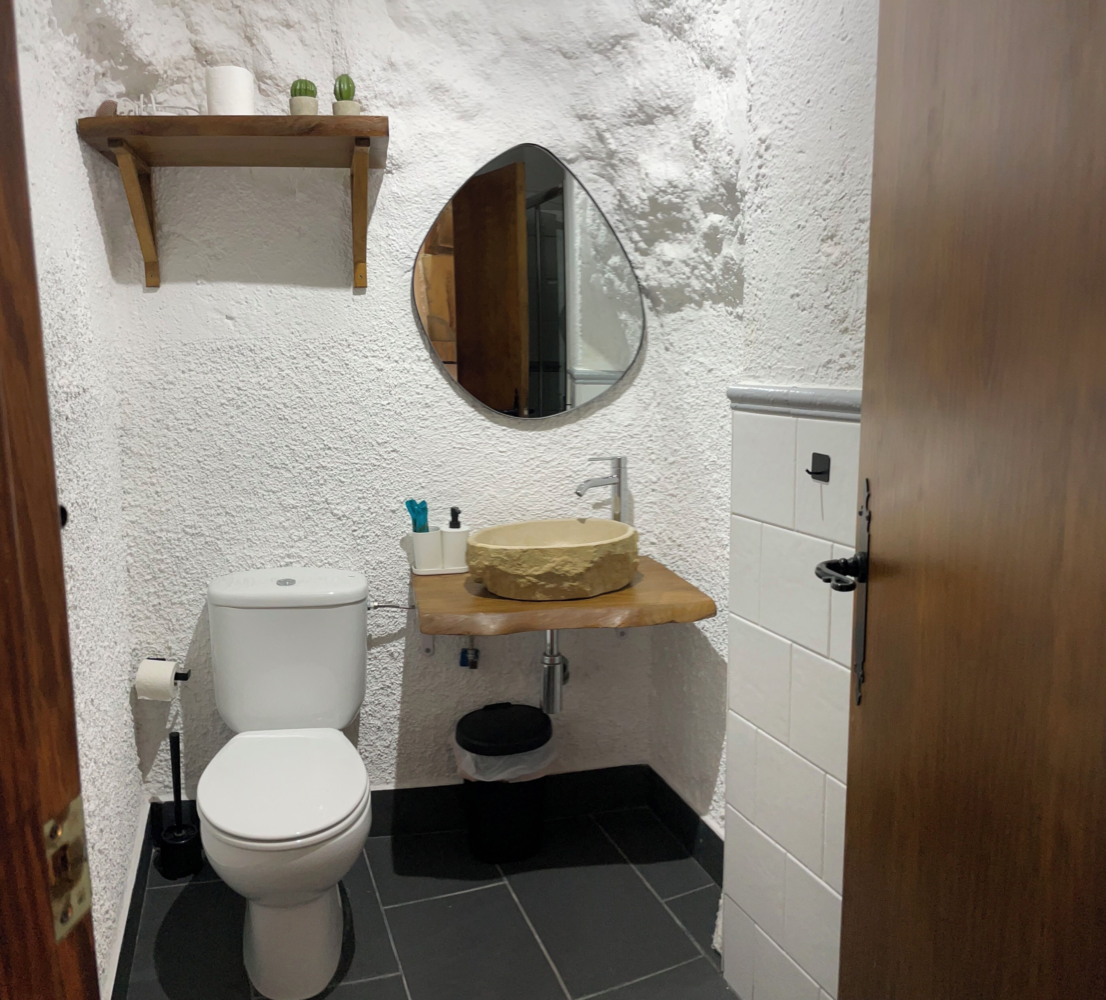 Un baño pequeño con inodoro, lavabo de encimera de madera, espejo y un estante con artículos de aseo.
