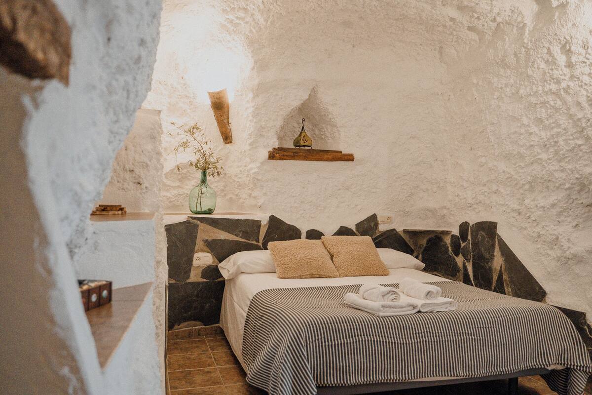 A cozy bedroom with a bed, pillows, a bedside table, a vase with flowers, and a shelf with a decorative item.