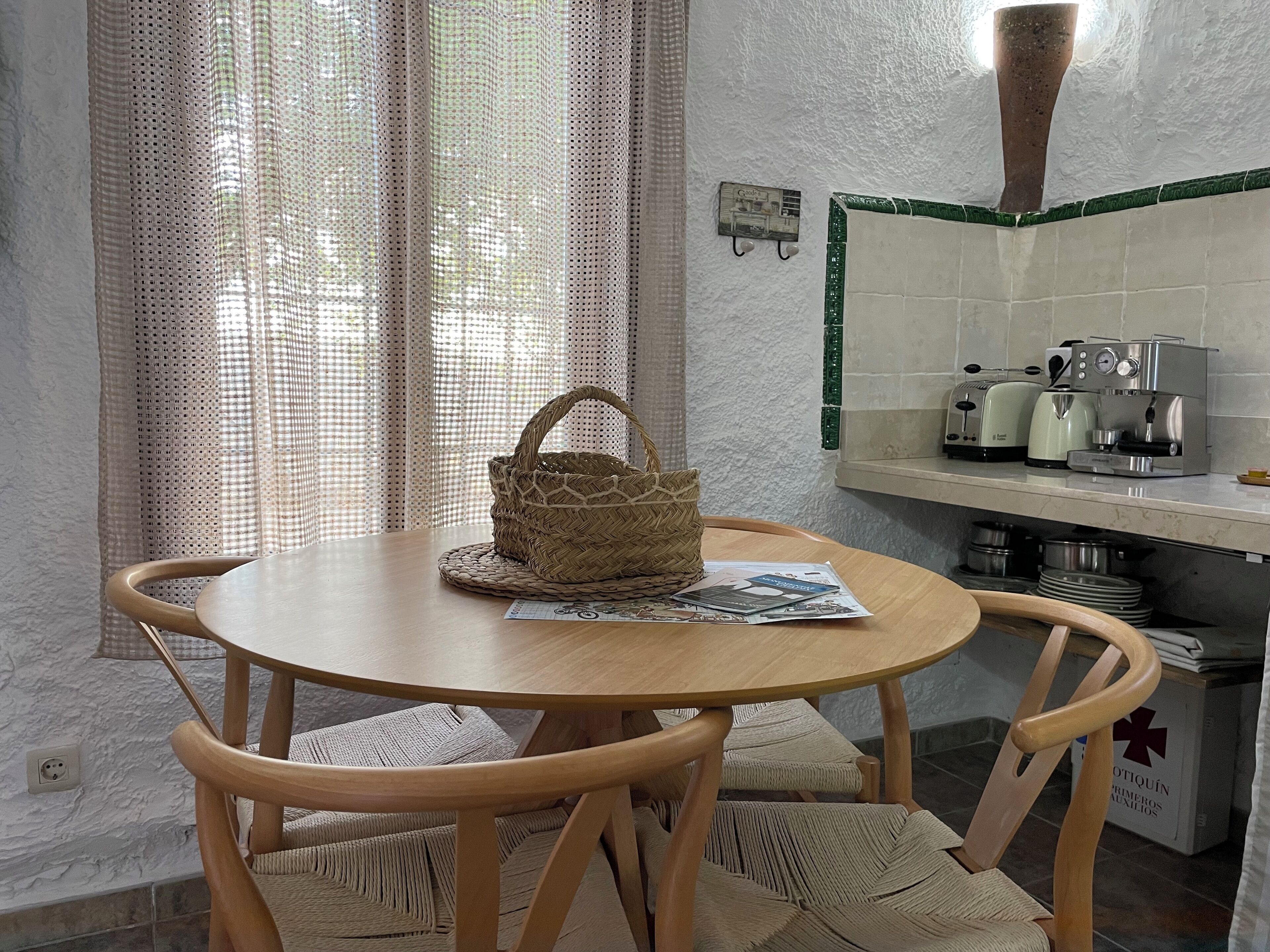 Una mesa redonda de madera con una cesta tejida y un periódico. Hay dos sillas de mimbre con cojines. Se ve una cocina con una cafetera, un tostador y ollas.