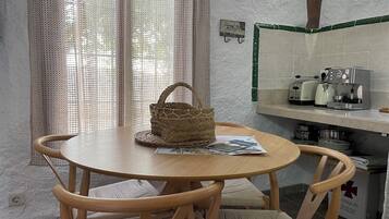 Una mesa redonda de madera con una cesta tejida y un periódico. Hay dos sillas de mimbre con cojines. Se ve una cocina con una cafetera, un tostador y ollas.