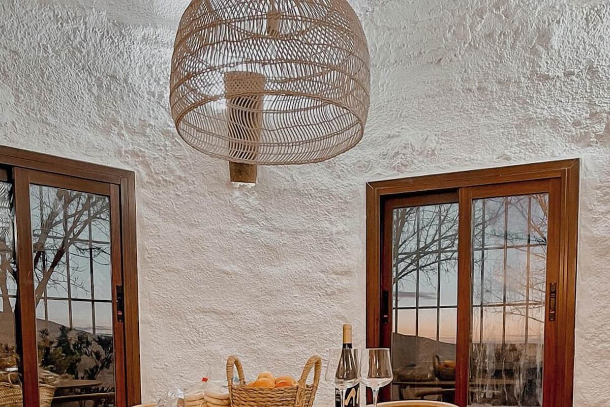 A round wooden table with woven chairs, a woven light fixture, and a view through large windows.