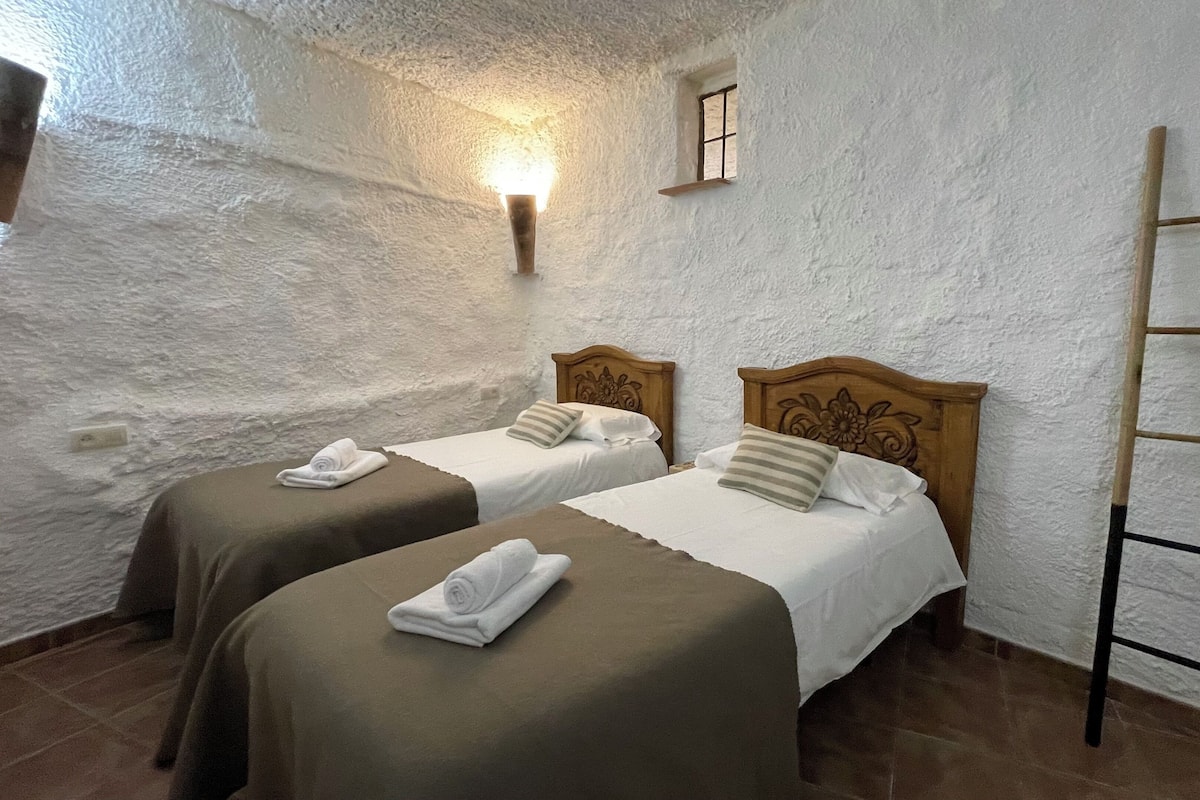 A room with two beds, each with a wooden headboard and white bedding. There is a small window with a wooden shelf above it, and a ladder leaning against the wall.
