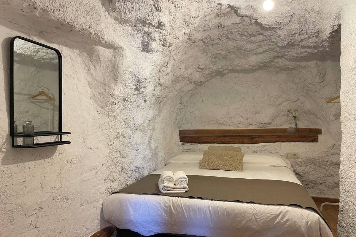 A rustic room with a bed, a wooden headboard, a mirror, and a shelf with a towel and a small vase.