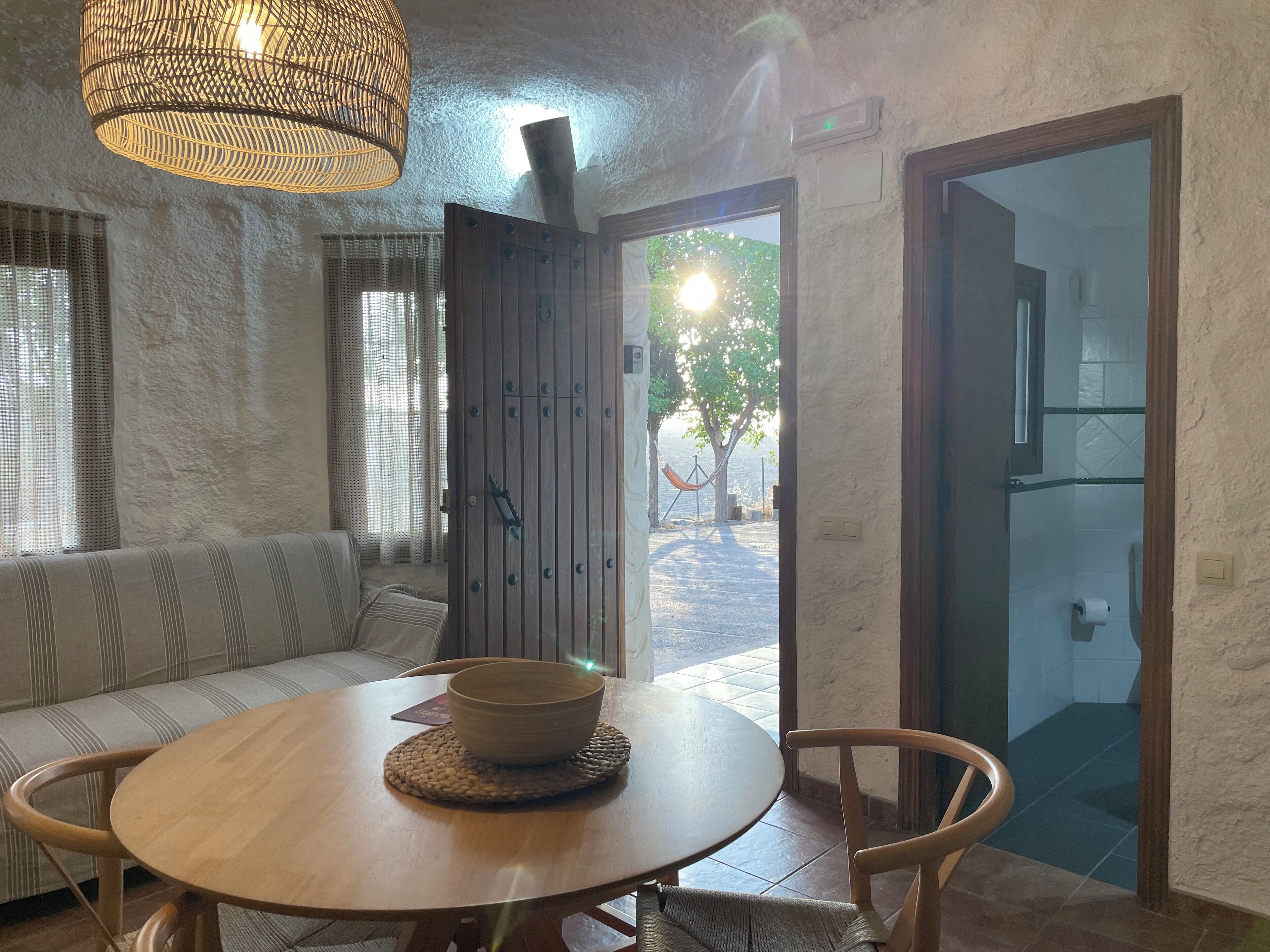 Une pièce chaleureuse avec une table ronde en bois, une chaise en osier, un banc rembourré et une grande porte donnant sur l’extérieur.