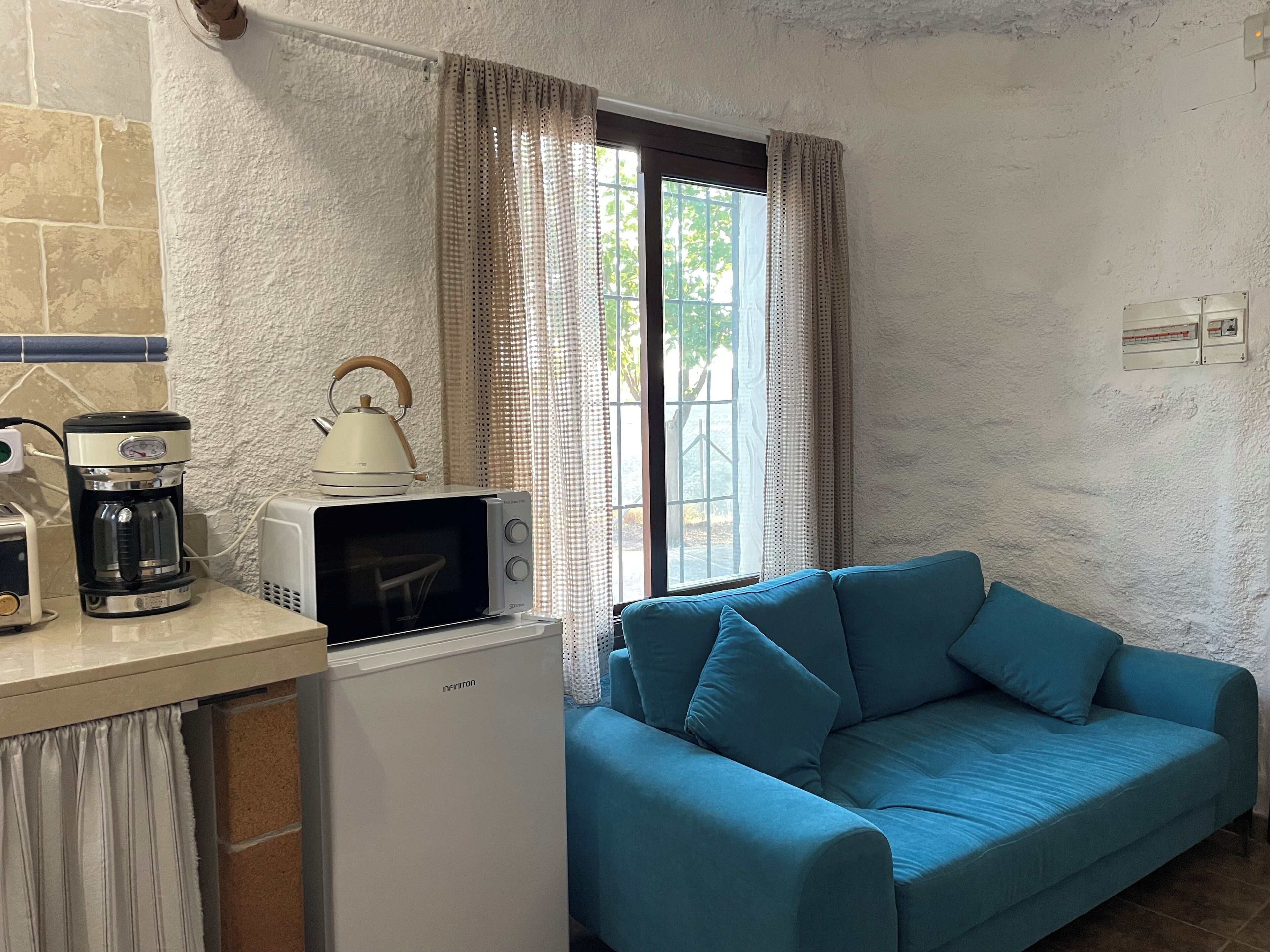 A kitchenette with a coffee maker, microwave, and refrigerator. There is a blue sofa and a window with curtains.