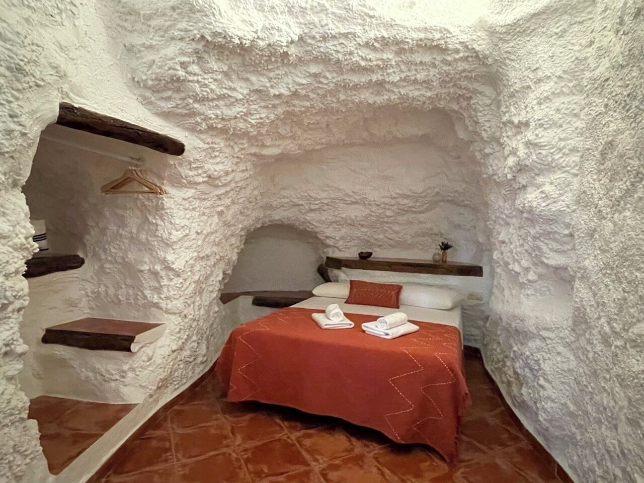 A small, rustic room with a bed, a wooden shelf, and a hanging garment rack.