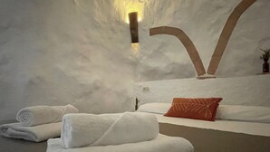 Habitación de spa con una pared grande, suave, blanca y texturizada, una cama con sábanas blancas y una almohada naranja.