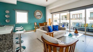 Een moderne woonkamer met een turquoise accentmuur, een plafondventilator, een bank met opvallende kussens, een salontafel en uitzicht op het balkon.
