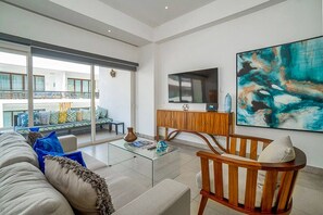 En moderne stue med sofa, sofabord og træstol. Der er et stort maleri på væggen og et fjernsyn på et træskab.