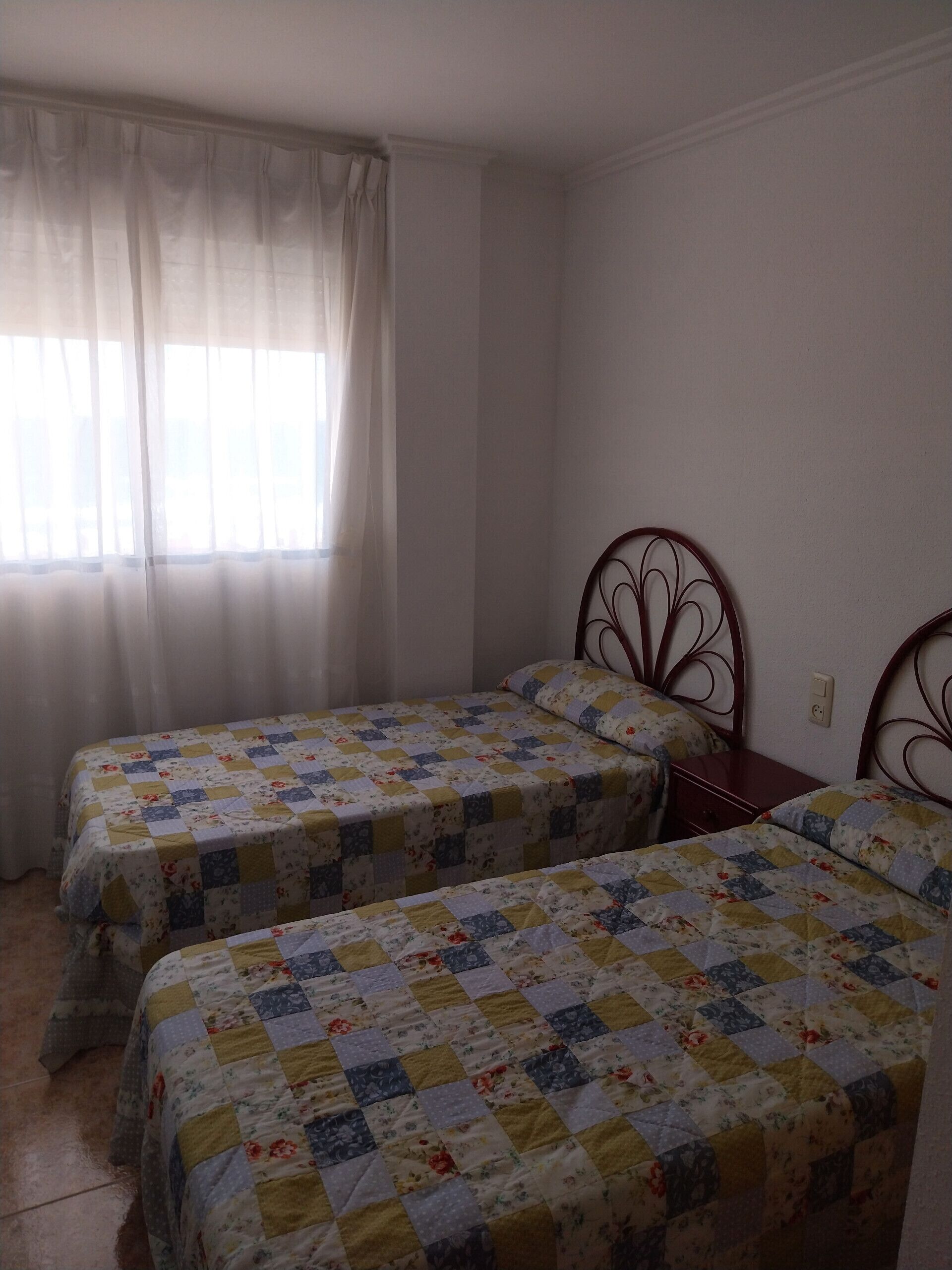 Quarto com duas camas de solteiro, cada uma com colcha floral, cabeceira de madeira e janela com cortinas leves.
