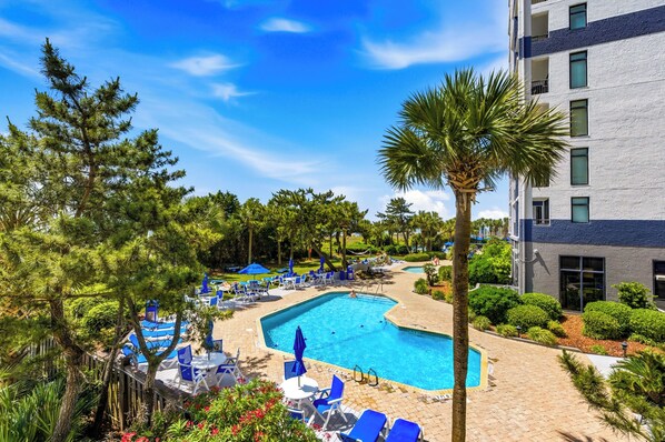 A pool area with lounge chairs and umbrellas, surrounded by trees and a building.