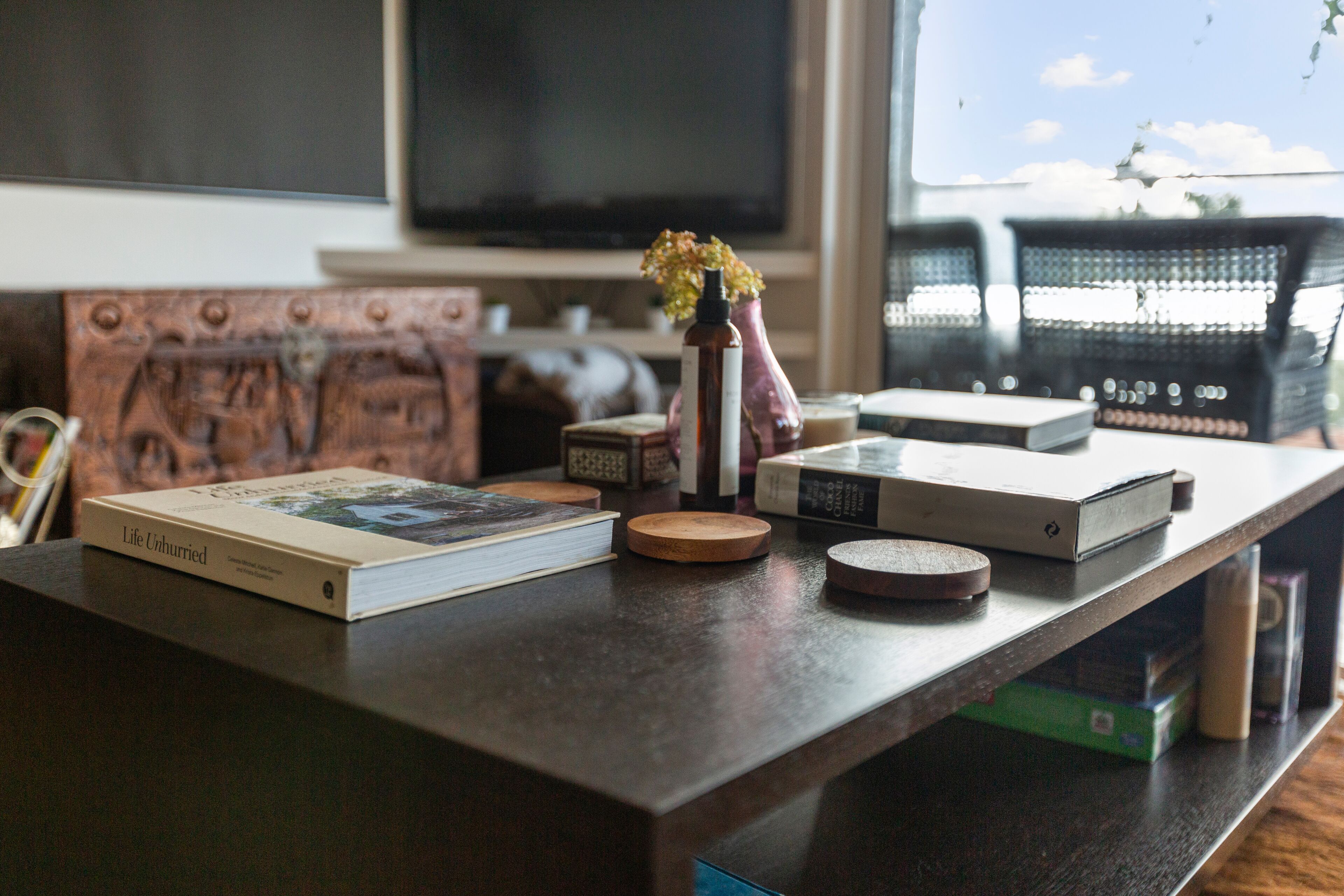 A wooden table with a book titled "Life Unhurried," a vase with flowers, and several round wooden coasters.