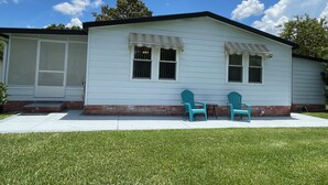 A single-story house with a front porch, two windows with awnings, and two green Adirondack chairs.
