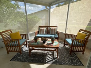 A sunlit patio with a wooden table and chairs, a patterned rug, and a view of a house and trees.