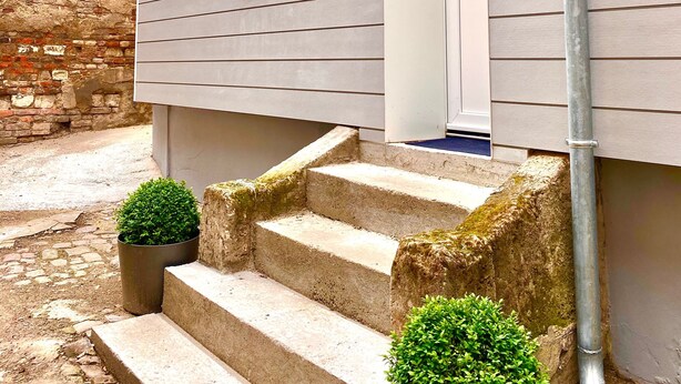 A building with a grey exterior, a set of concrete steps leading to a white door, and a small potted plant at the bottom of the steps.