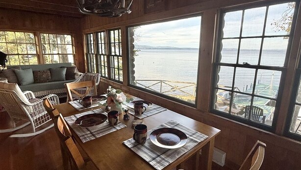A wooden dining area with a table set for four, large windows overlooking a body of water, and a cozy seating area with a sofa and chairs.