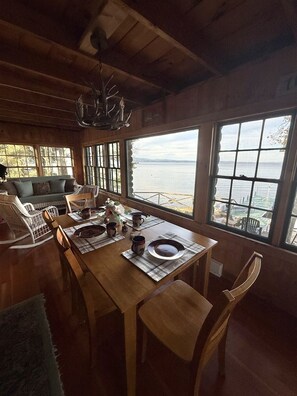 A wooden dining area with a table set for four, large windows overlooking a body of water, and a cozy seating area with a sofa and chairs. - Waterfront on Sebago Lake (Standish)