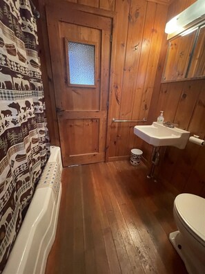 Bathroom features a white bathtub, sink, and toilet, set amidst rustic wooden walls and a charming door with a small window.