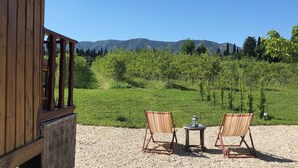 A wooden building with a balcony overlooking a vineyard and mountains. There are two chairs and a table with a lantern on the balcony. - Roulotte 2 Personnes (Saint-Rémy-de-Provence)