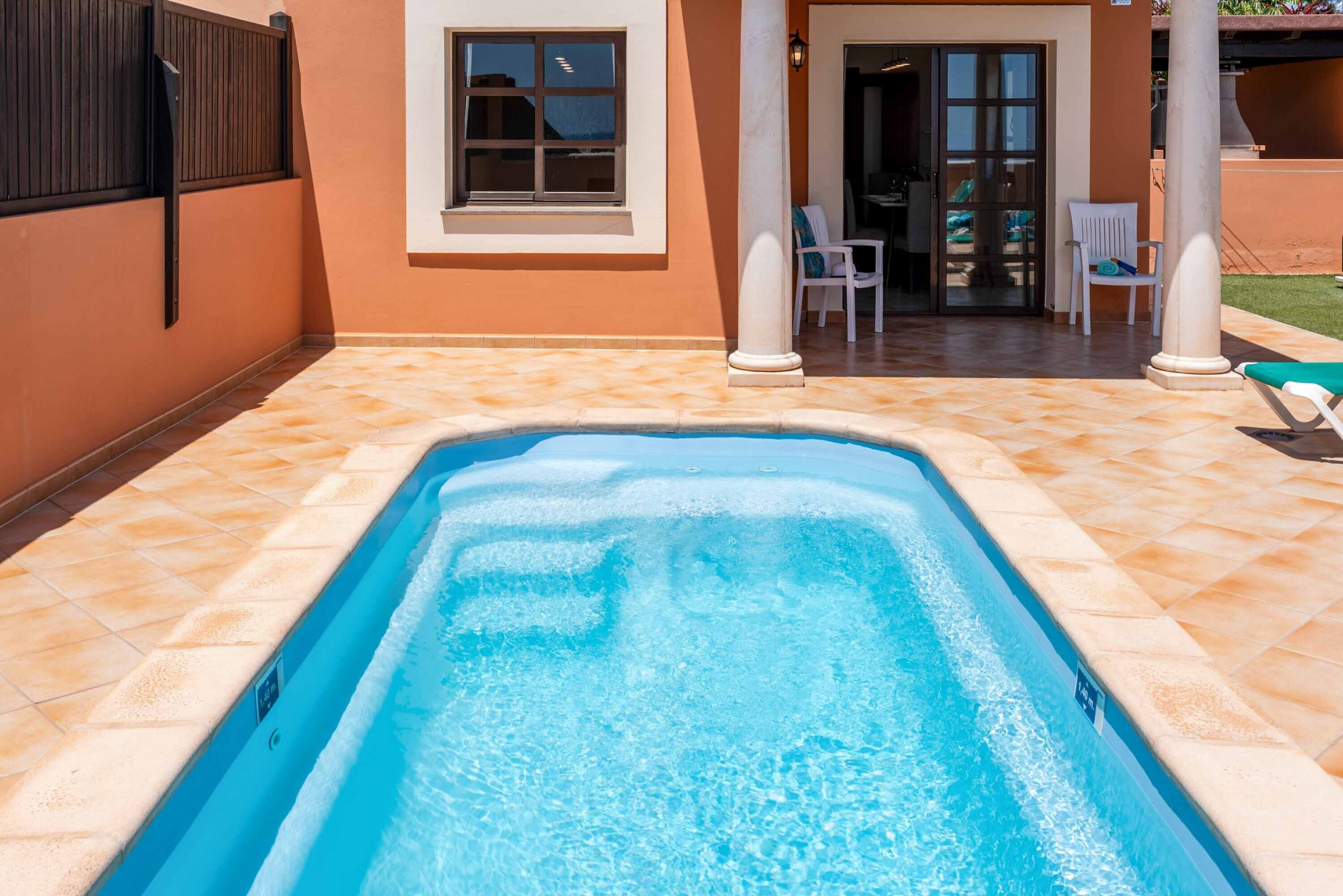 Ein rechteckiger Swimmingpool mit klarem, blauem Wasser, umgeben von beigefarbenen Fliesen und einer weißen Wand.