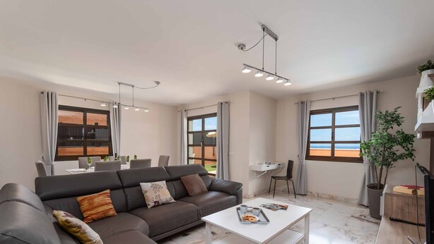 Ein modernes Wohnzimmer mit einer Eckcouch, einem weißen Couchtisch und einem Essbereich mit Tisch und Stühlen. Große Fenster mit Vorhängen, eine Deckenleuchte und ein Topf mit Pflanze sind ebenfalls vorhanden.