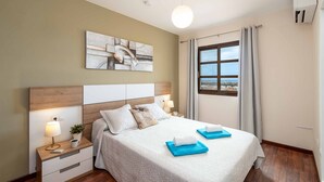 Ein Hotelzimmer mit Bett, Nachttischen, einer Lampe, einem Gemälde, einem Fenster mit Vorhängen und einer Klimaanlage.