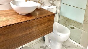 A bathroom with a marble-tiled wall, a wooden vanity, a white sink with a gold faucet, a round mirror, a glass shower enclosure, and a toilet. - Appartement REZ DE Chaussee (Amiens)