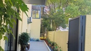 Un étroit sentier mène à un bâtiment dont l’entrée est protégée par un portail, et qui est entouré de plantes en pot et d’un petit jardin.