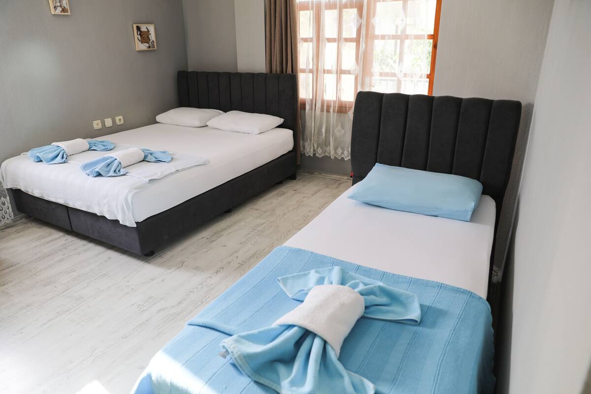 A hotel room with two beds, a large window with sheer curtains, and a light-colored wooden floor.