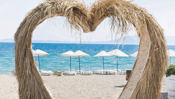 Una playa con un arco de paja con forma de corazón, sillas de descanso blancas y sombrillas.