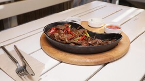 Plato servido en una sartén negra sobre una tabla de madera, acompañado de un tenedor y servilleta sobre una mesa blanca.