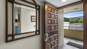 Puerta de madera con espejo y cerradura electrónica, que da a un balcón con vistas a la vegetación y un paisaje lejano.