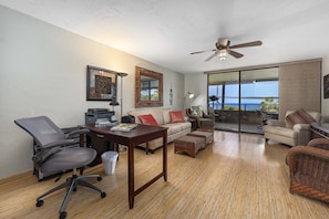 Uma sala de estar espaçosa com piso de madeira, um sofá, uma mesa de centro, uma escrivaninha com cadeira e uma grande janela com vista para o oceano.