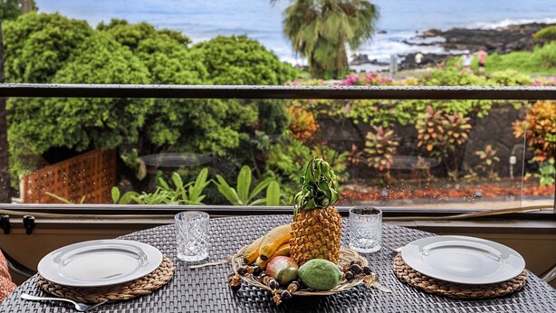 Una mesa con un posavasos tejido, una piña, un mango y una fruta verde, con vista al océano y follaje en el fondo.